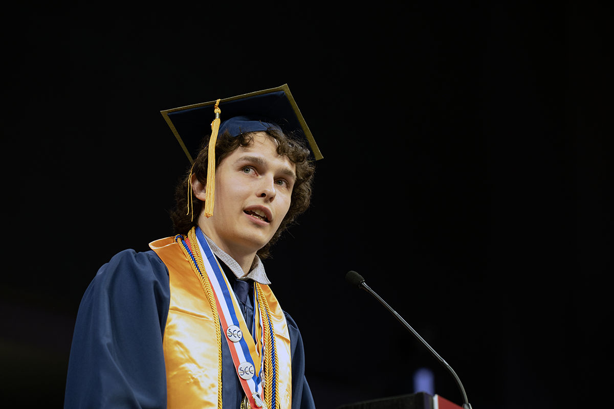Adam Long in cap, gown and honors standing at podium speaking at Scott's Commencement Ceremony