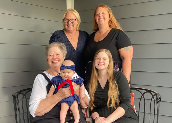 Five generations of women in the same family smile for a photo on a front porch. Seated in front, Haylea Mallia and her great-grandmother Leota Rickey hold Haylea’s baby daughter, Lily, who is dressed in a blue sailor-style outfit with a red ribbon. Behind them stand Haylea’s mother, Heather Scranton, and grandmother. All are smiling and gathered closely together.