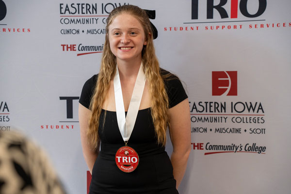 Haylea Mallia stands in front of a step-and-repeat banner at the 2025 TRIO Graduation Night. She wears a black dress and a TRIO Graduate medal around her neck, smiling proudly.