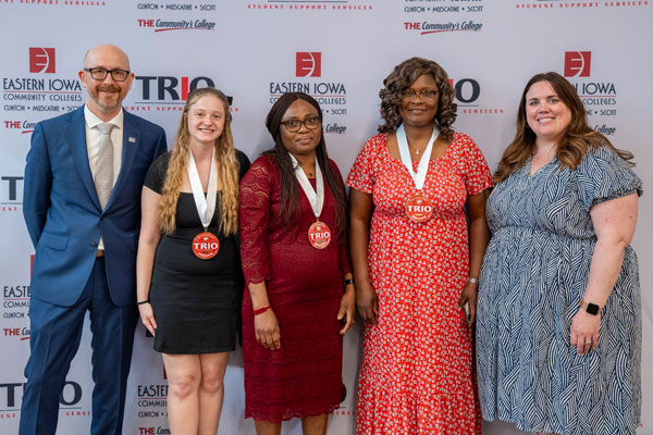 Haylea Mallia stands with fellow TRIO graduates and staff members in front of a step-and-repeat banner at the 2025 TRIO Graduation Night. She wears a black dress and a TRIO Graduate medal around her neck, smiling proudly alongside her peers and program supporters.