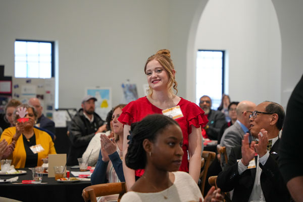 Haylea Mallia, wearing a red short-sleeve blouse, stands up for recognition during the 2025 SCC Student Showcase event. Seated attendees are clapping for her. 