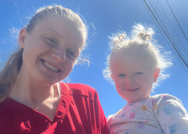 Haylea Mallia, wearing red nursing scrubs, smiles alongside her young daughter, Lily, under a bright blue sky.